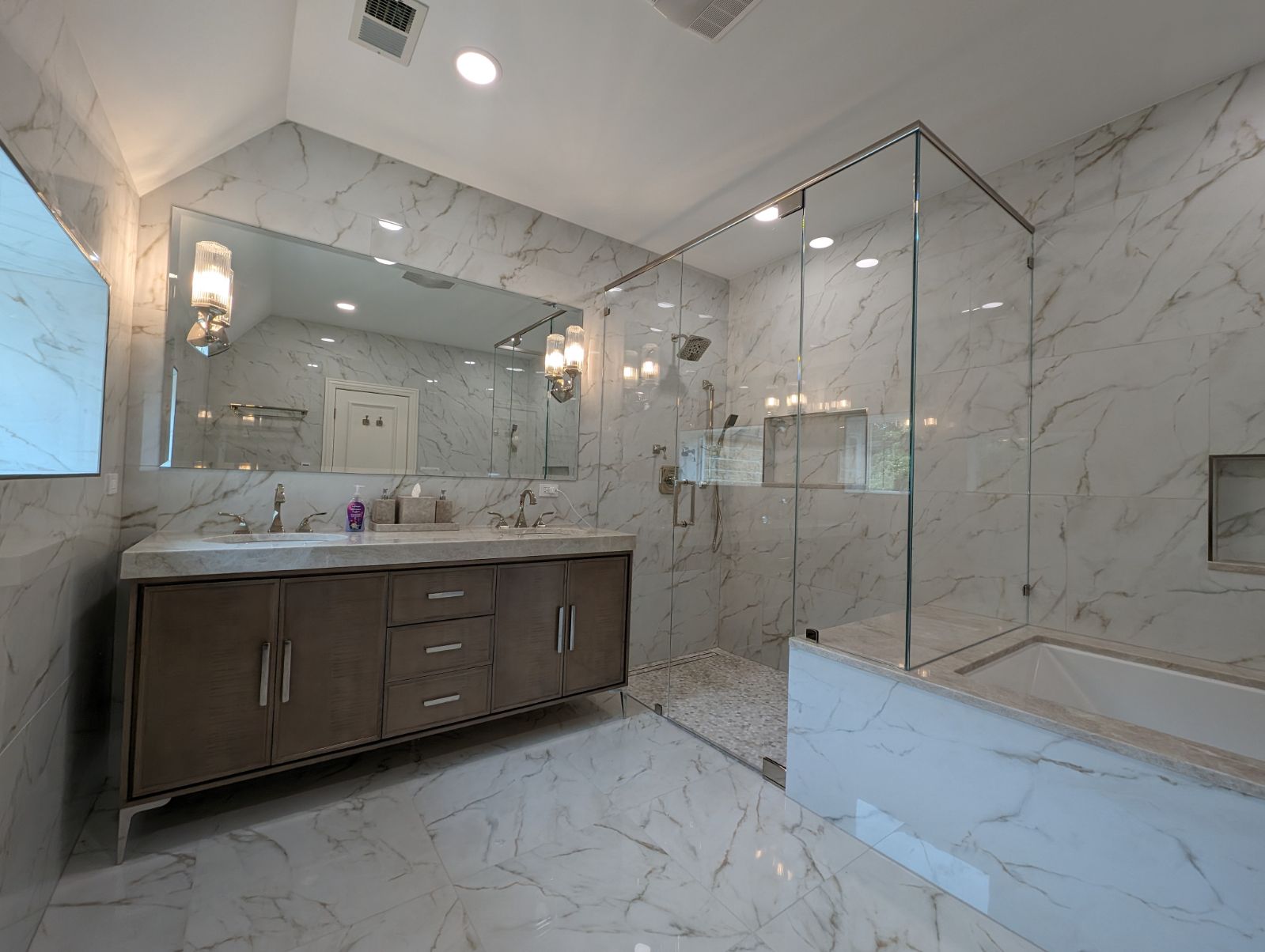 An expansive bathroom crafted in white marble, ashy wood, and glass. A vanity and shower stand against one wall while a bathtub stands against another.